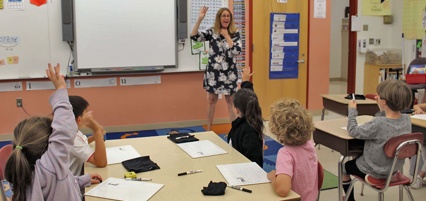 A teacher in a floral dress stands at the front of a classroom with a whiteboard, engaging with young students who are seated at desks and raising their hands. Various educational materials are displayed on the walls.