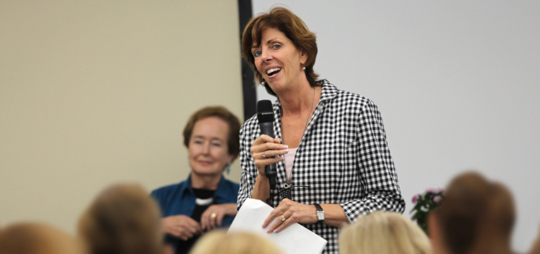 A woman in a checkered shirt speaks into a microphone, smiling and holding papers. Another woman stands in the background, partially in view, in a blue and black outfit. Both are in a room with a blurred audience in the foreground.