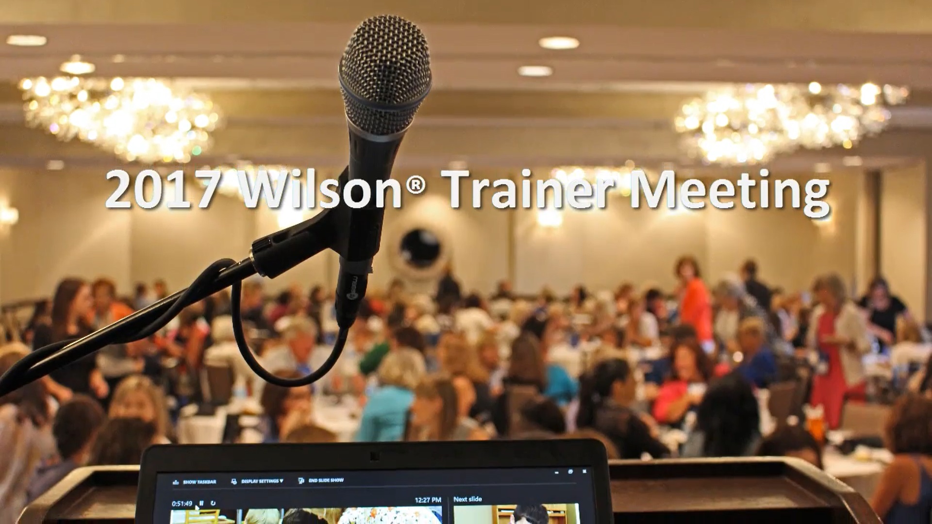 A microphone on a stand is in focus in a conference room setting. In the background, a large crowd of people sit at tables. Text on the image reads 2017 Wilson® Trainer Meeting.