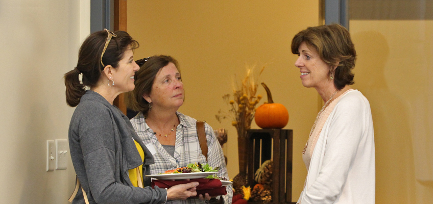 Three women are standing and talking in a room. One woman is holding a plate with a salad. There is a pumpkin and autumn decorations on a shelf in the background.