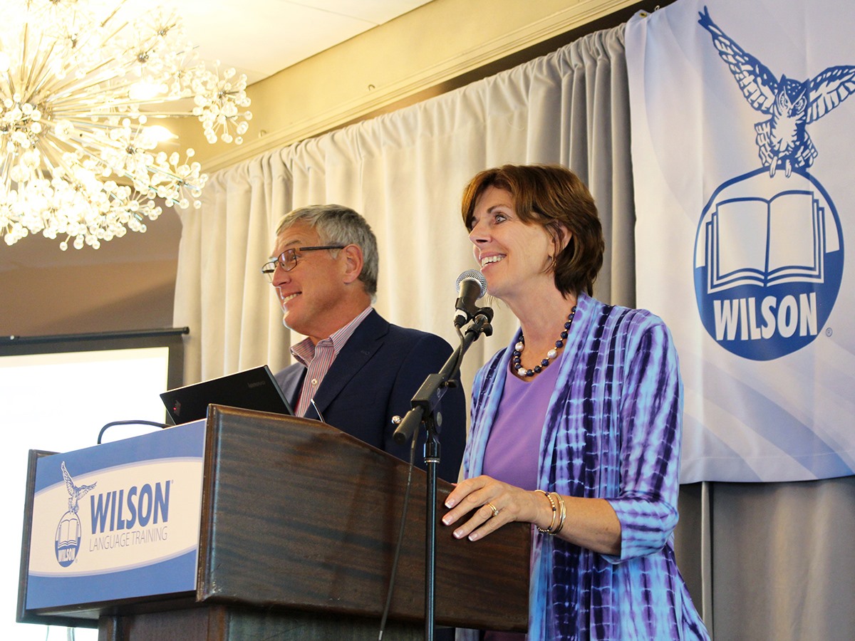 A man and a woman stand smiling behind a podium with Wilson Language Training signage. The woman speaks into a microphone while the man holds a tablet. A blue banner with an owl logo and Wilson text is displayed behind them.