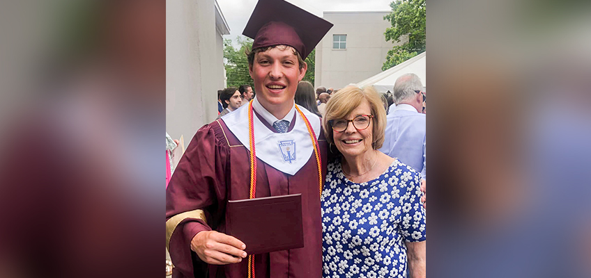 A graduate in a maroon cap and gown smiles while holding a diploma. He stands beside an elderly woman in glasses, wearing a blue floral dress. A building and people are visible in the background.