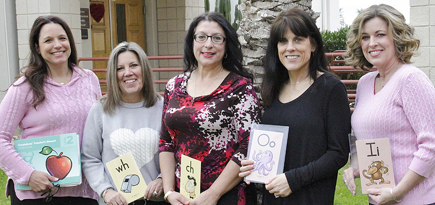 Five women smiling and holding educational materials with letter and image cards in front of a building.