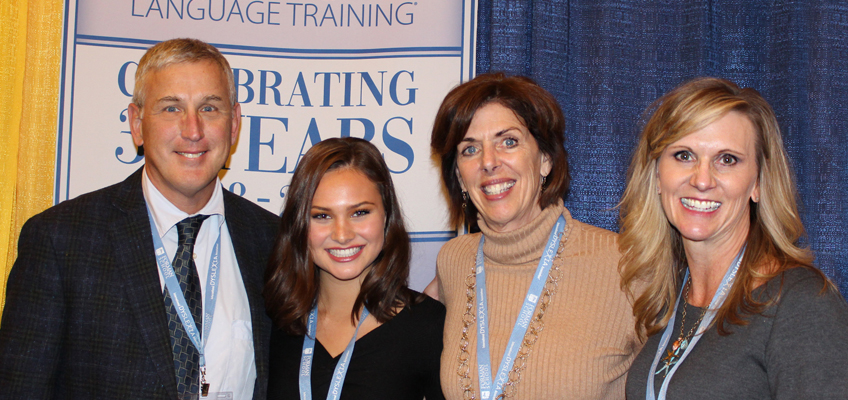 Four people smile for a group photo at an event, standing in front of a banner that mentions a 30-year celebration related to language training. They are wearing name tags and lanyards, indicating their participation in the event.
