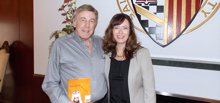 A man and woman, both smiling, stand in front of a coat of arms. The man is holding an orange book. They are indoors, near a chair and some plants.