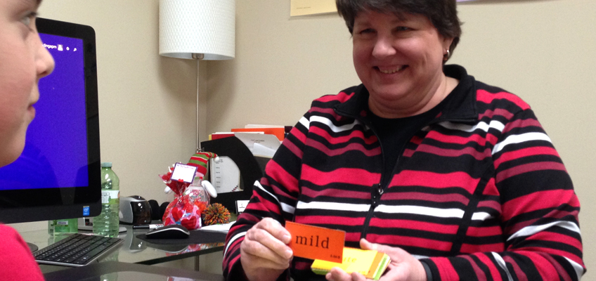 A woman wearing a striped sweater holds a flashcard while sitting at a desk. She is smiling at a child seated across from her. A computer monitor and various office supplies are visible in the background.