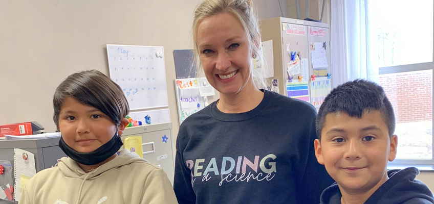 A woman smiling in a classroom, standing between two children. The woman wears a black shirt with Reading a Science printed on it. The children, one wearing a mask around their neck, are smiling. Classroom materials and a window are in the background.
