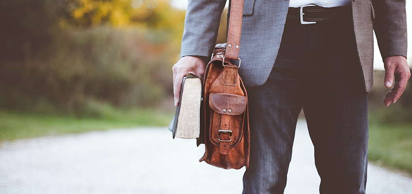 A person wearing a gray blazer and jeans is standing outdoors, holding an old book and a brown leather bag. The background is slightly blurred, showing a pathway and green foliage.