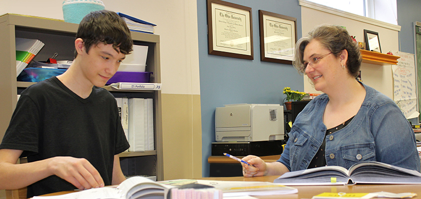 A woman and a teenage boy are sitting at a desk in an office, engaged in conversation. The woman is holding a pen and smiling, while the boy looks at a book. Shelves, a printer, and framed certificates are visible in the background.