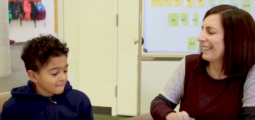 A woman and a young boy sit together in a classroom setting. The woman is smiling and appears to be engaged in conversation with the boy, who is looking down with a slight smile. A whiteboard and colorful backpacks are visible in the background.