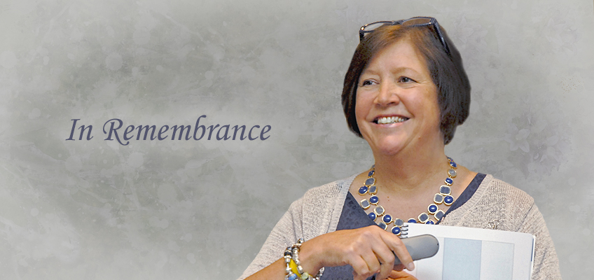 Smiling woman holding a document and remote, wearing a blue necklace, with glasses on her head, against a gray background with the text In Remembrance.