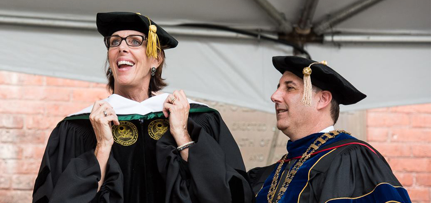 A person in a graduation cap and gown joyfully holds two medallions around their neck. Beside them, another person in similar attire smiles. They are outdoors under a canopy with a brick wall in the background.