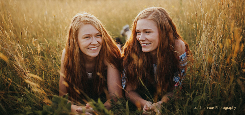 Two people with long red hair lie on their stomachs in a grassy field, smiling warmly. The sunlight casts a golden glow over the scene, creating a serene and cheerful atmosphere.