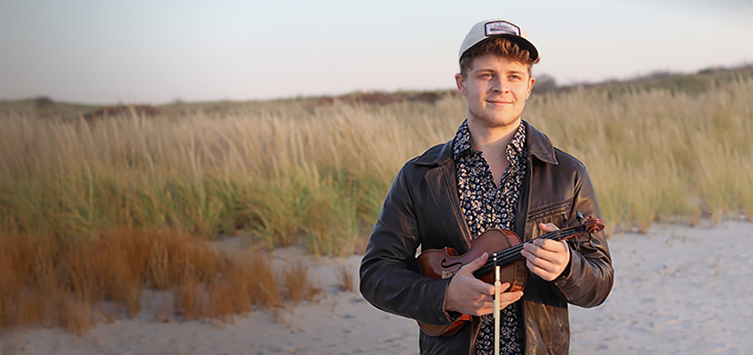 A person stands on a sandy beach, holding a violin. They are wearing a floral shirt, a dark leather jacket, and a cap. The background features tall grasses and a clear sky, with soft lighting suggesting early morning or late afternoon.