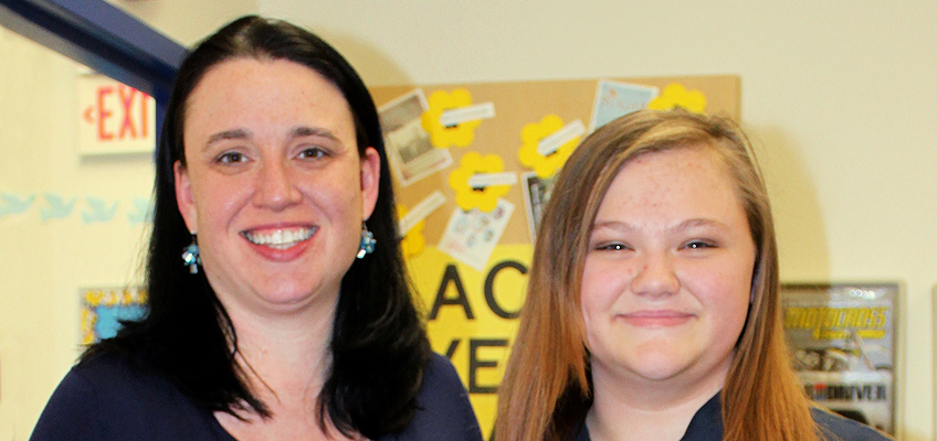 Two people smiling in front of a bulletin board covered with papers and decorations. The person on the left has long dark hair, and the person on the right has long light brown hair.