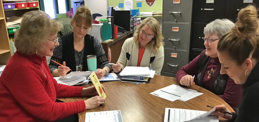 A group of five women sitting around a table engaged in discussion. They have papers and pens, indicating a meeting or collaborative work environment. Books and file cabinets are visible in the background.