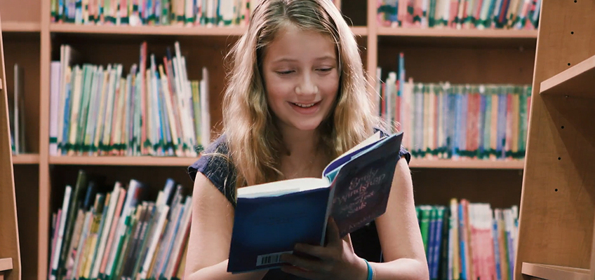 A young girl with long blond hair is sitting and smiling while reading a book in a library. Bookshelves filled with various colorful books are visible in the background.