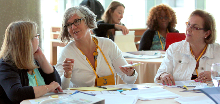 A group of women are sitting around a table with papers and folders. One woman with glasses and short gray hair is speaking, holding papers. Others are listening attentively. Some are wearing yellow lanyards. The setting appears to be a conference or meeting room.