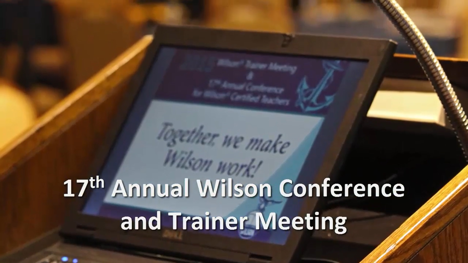 A laptop on a podium displays a presentation slide with the text Together, we make Wilson work! Below, it reads 17th Annual Wilson Conference and Trainer Meeting. The background is blurred, focusing on the laptop screen.