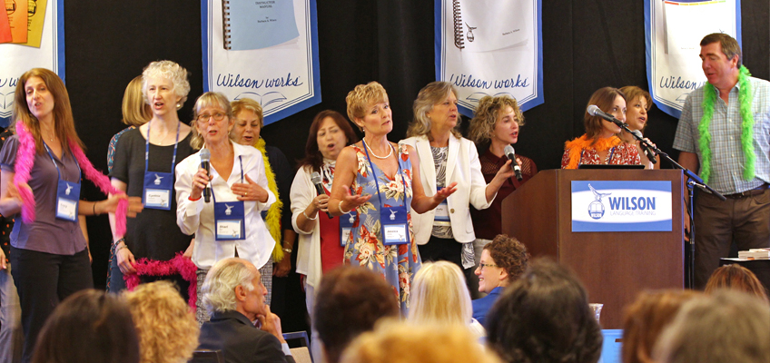 A group of people stand on a stage, some singing and gesturing expressively. They wear various colorful clothing and name tags. A podium with a microphone is to the right, and banners with Wilson logos hang in the background. An audience is visible.