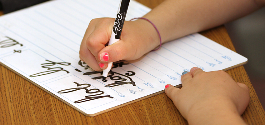 A child practices cursive writing on a whiteboard with lines. The child uses a black marker to trace the words. The board rests on a wooden table, and the child wears a pink bracelet.