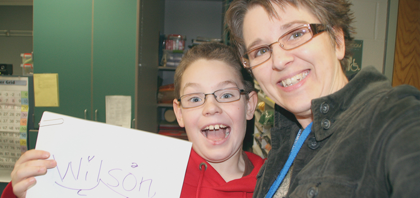 A smiling adult with short hair and glasses takes a selfie with a child wearing glasses and a red hoodie. The child holds up a piece of paper with Wilson written on it. They are in a room with green cupboards and various items in the background.
