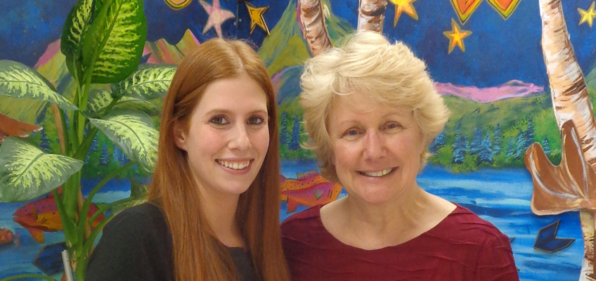 Two women smiling at the camera. The woman on the left has long red hair and wears a black top. The woman on the right has short blonde hair and wears a red top. They stand in front of a colorful, tropical-themed mural.
