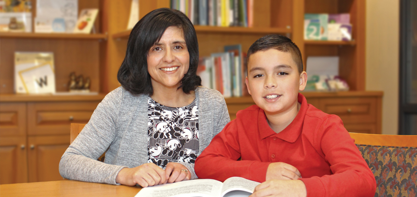 A woman and a boy sit at a table, smiling at the camera. A book is open in front of them. Bookshelves filled with books are in the background. The woman wears a patterned blouse and cardigan, and the boy wears a red shirt.