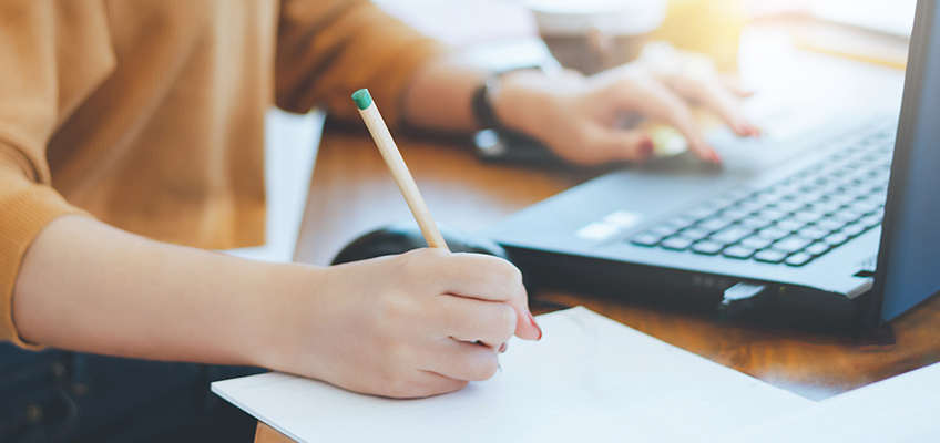 A person in a sweater is seated at a wooden desk. They are using a laptop with one hand while holding a pencil and writing in a notebook with the other. The workspace is brightly lit and tidy.