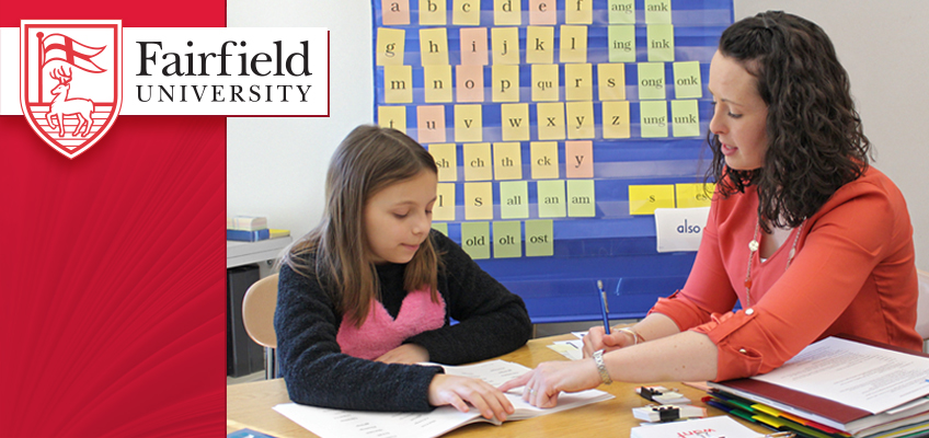 A teacher sits at a table with a young student, pointing to a book as they read together. Behind them is a wall display with letters and words. The Fairfield University logo is in the upper left corner.
