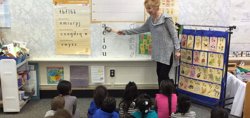 A teacher stands at the front of a classroom, pointing at the vowels a e i o u on a board. Several young children sit on the floor, attentively watching. The classroom walls are adorned with educational posters and charts.