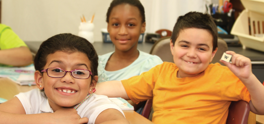 Three smiling children in a classroom, sitting at a table. The child on the left wears glasses, the one in the middle is in a blue shirt, and the one on the right, in an orange shirt, holds a wooden block with a W. Books and supplies are visible in the background.