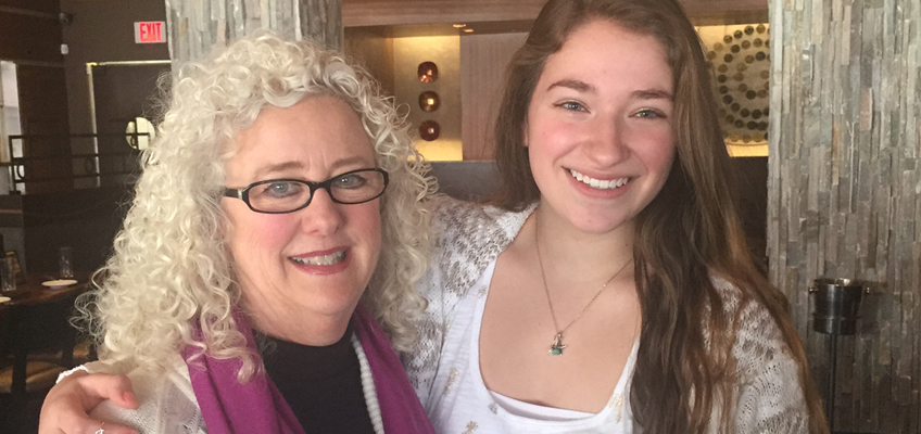 Two women smiling indoors. The older woman has curly white hair, glasses, and is wearing a purple scarf over a black top. The younger woman has long brown hair and is wearing a white top with necklaces. They are standing close together in a warmly lit room.