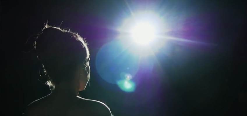 A silhouette of a person with their hair in a bun is facing a bright, white spotlight. The light creates lens flare effects, and the background is dark, emphasizing the light's intensity.