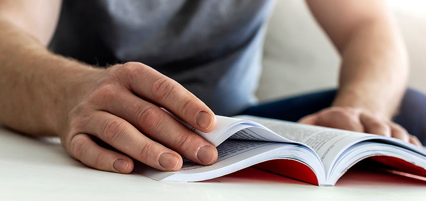 A person is sitting and flipping through a book with a red interior cover. The focus is on the hand turning a page. The background is blurred, and the person's torso is partially visible, wearing a gray shirt.