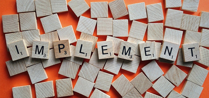 Wooden Scrabble tiles spell out IMPLEMENT on an orange background. Surrounding the word are several blank wooden tiles.