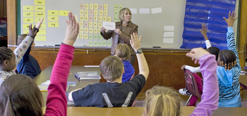 A teacher stands in front of a classroom holding a flashcard, while several students raise their hands. The board behind the teacher displays various word cards. Students are seated at desks facing the teacher.