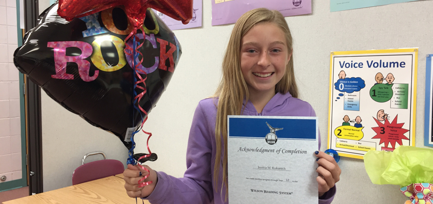 Girl smiling and holding a certificate in one hand and a black star-shaped balloon with 100 ROCK in colorful letters in the other. She stands in a classroom with educational posters on the wall.