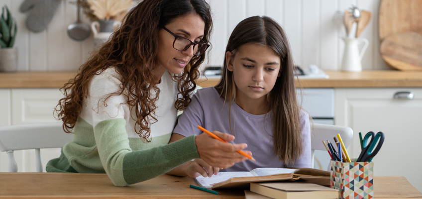 A woman with long curly hair and glasses helps a young girl with long straight hair study at a table. They are surrounded by books and stationery in a cozy kitchen setting. The woman is pointing at a notebook with a pencil.