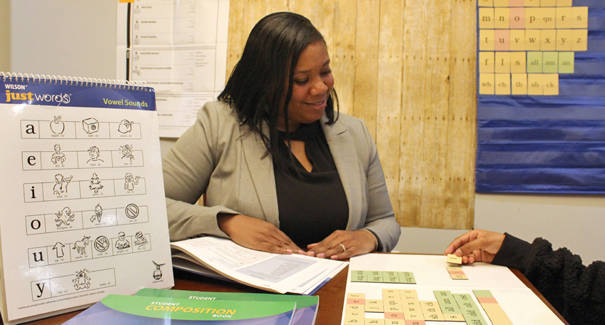A teacher sits at a table with a student, engaging in a phonics lesson. Educational materials with letters and sounds are spread out, including a chart and a workbook. A poster with letters is on the wall. Both are focused on the activity.