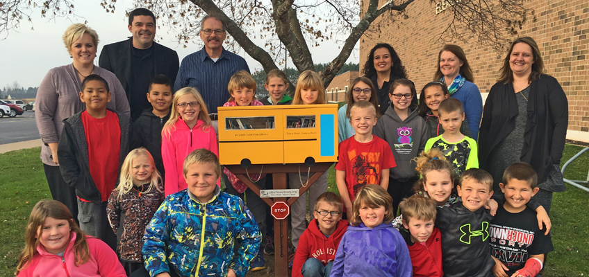 A group of young students and adults pose outdoors around a small yellow box resembling a school bus, mounted on a post. Some children are kneeling, and others stand beside the adults. Leafless tree branches are visible in the background.