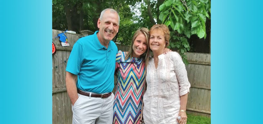 Three people smiling and standing closely together in a backyard. The man on the left is wearing a teal shirt, the woman in the middle has a colorful dress, and the woman on the right is in a light blouse. A wooden fence and green trees are in the background.