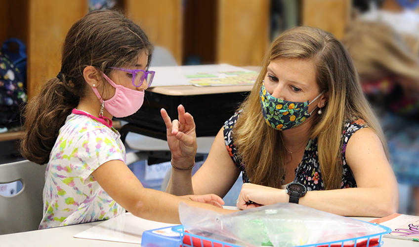 A young girl wearing glasses and a mask is sitting at a table with a woman, who is also masked. The woman is gesturing with her hand and smiling. The scene appears to be in a classroom or learning environment.
