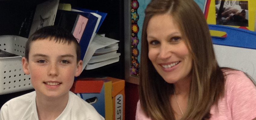 A woman and a boy sit closely, both smiling at the camera. They are indoors, with shelves containing books and colorful paper in the background. The woman has long brown hair, and the boy has short dark hair.