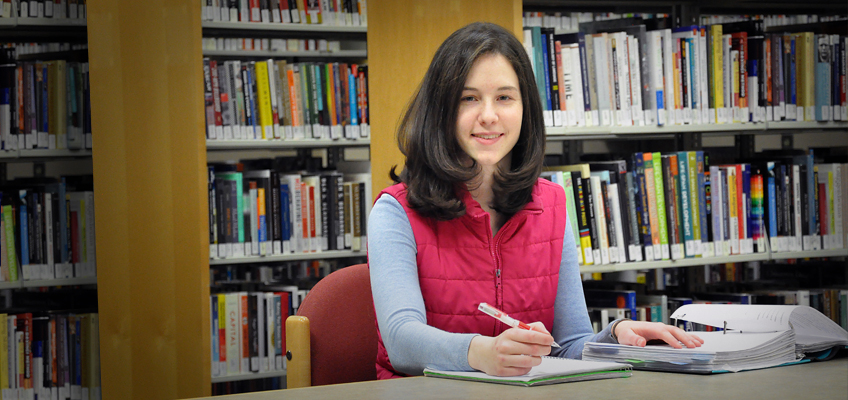 A person with long dark hair sits at a library table, smiling and holding a pen. They are wearing a red vest over a blue shirt. Open books and notebooks are spread out in front of them, with shelves of books in the background.