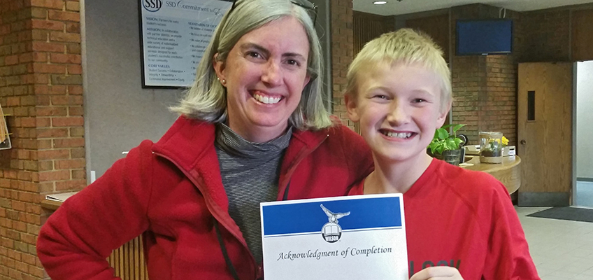 A woman and a smiling boy stand together indoors. The boy holds a certificate with a blue banner and an emblem. They both wear red tops and stand in a warmly lit room with brick walls and a counter in the background.