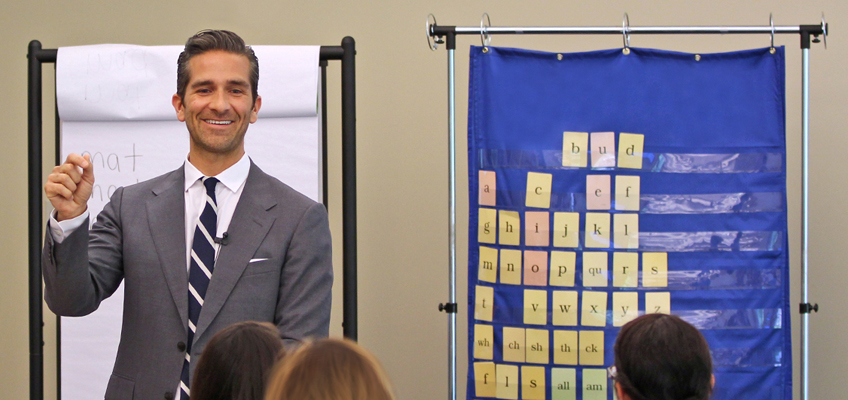A man in a suit is smiling and gesturing with his hand in front of an audience. Behind him are a flip chart and a blue pocket chart with alphabet cards and words. Several people are seated and facing him.