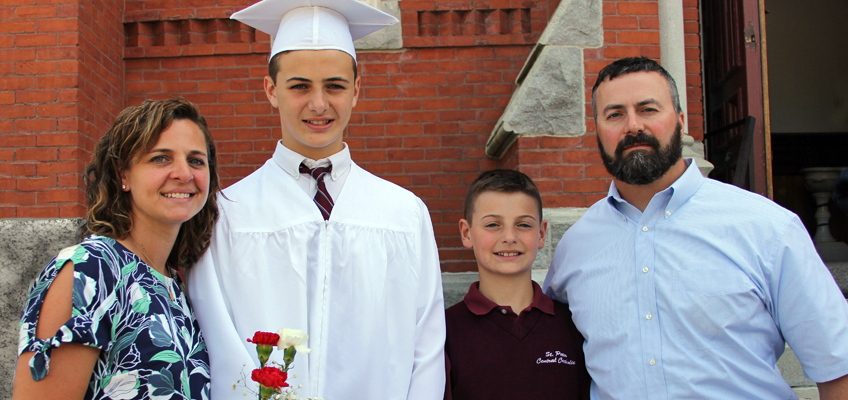 A smiling family stands together in front of a brick building. The person in the center wears a white graduation cap and gown, holding flowers. Two adults and a younger child stand beside them, all dressed in casual clothes.