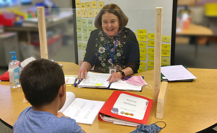 A teacher sits at a desk, smiling at a student across a clear divider. The table has books, papers, and sanitizer. The background has educational materials on the wall.
