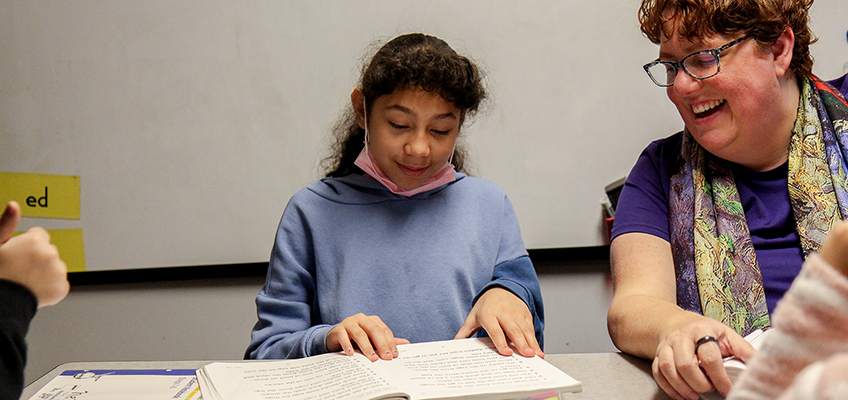 A young girl in a blue hoodie is reading a book at a table, with an adult woman wearing glasses and a colorful scarf sitting next to her, smiling. They appear to be in a classroom setting.